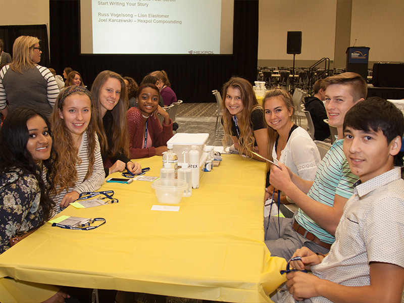 A group of smiling students sitting at a table, taking part in a demonstration at an indoor conference